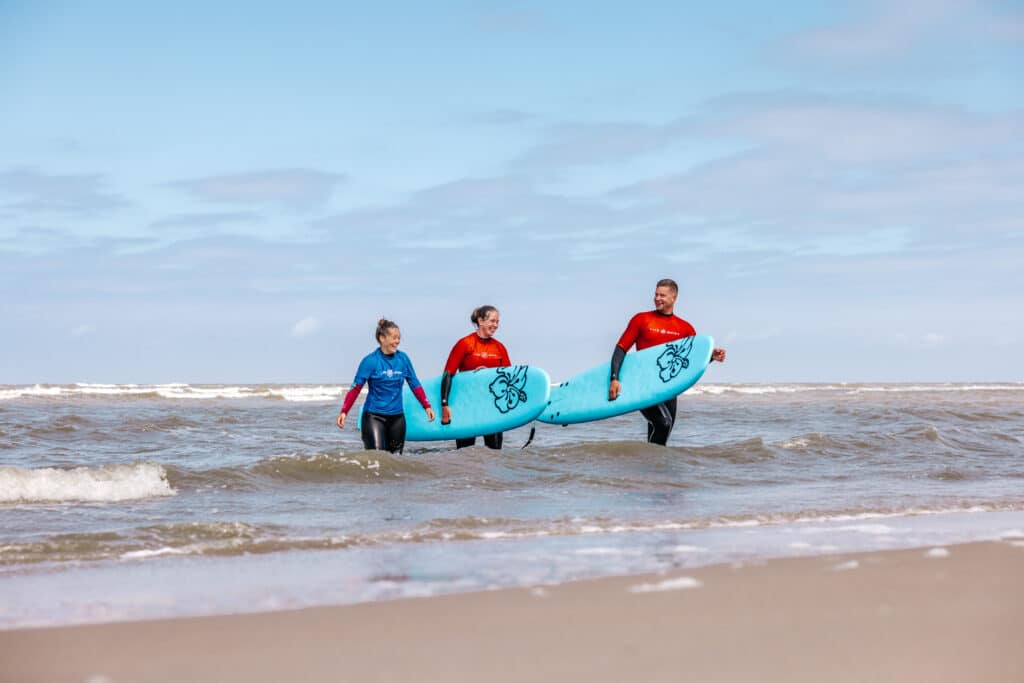 Een man en vrouw lopen na de surfles met hun surfplank uit de zee al pratend met hun surf instructrice