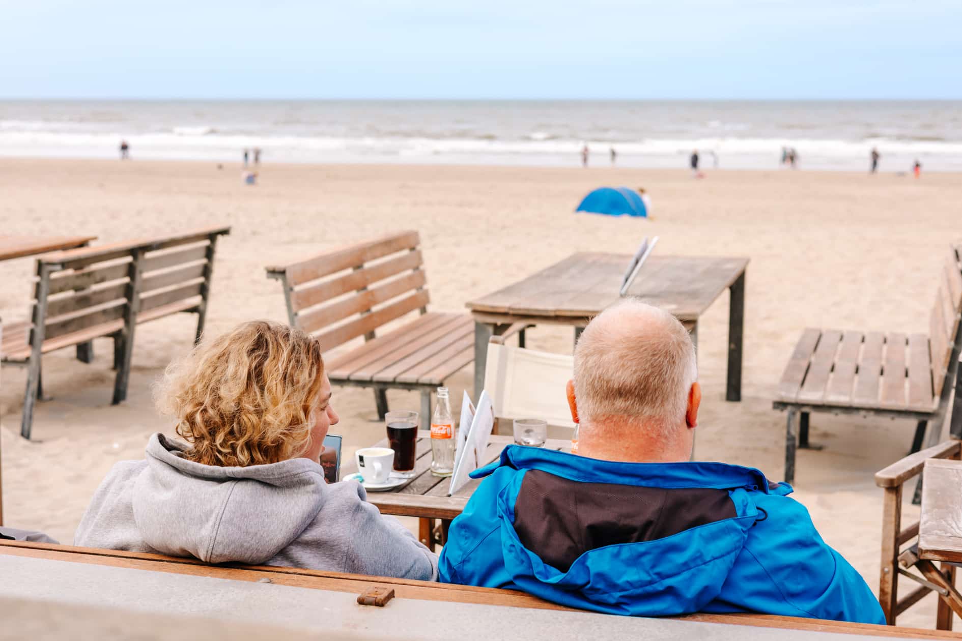 Twee mensen in oktober op het terras bij jaarrond strandpaviljoen De Haven van Zandvoort, uitkijkend over het strand en de zee