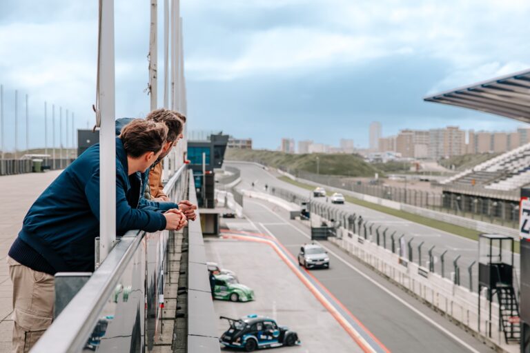 Een groep vrienden kijkt vanaf het uitzichtpunt op het circuit naar de baan, waar auto's aan komen rijden, met de hoofdtribune op de achtergrond. Sporten in Zandvoort