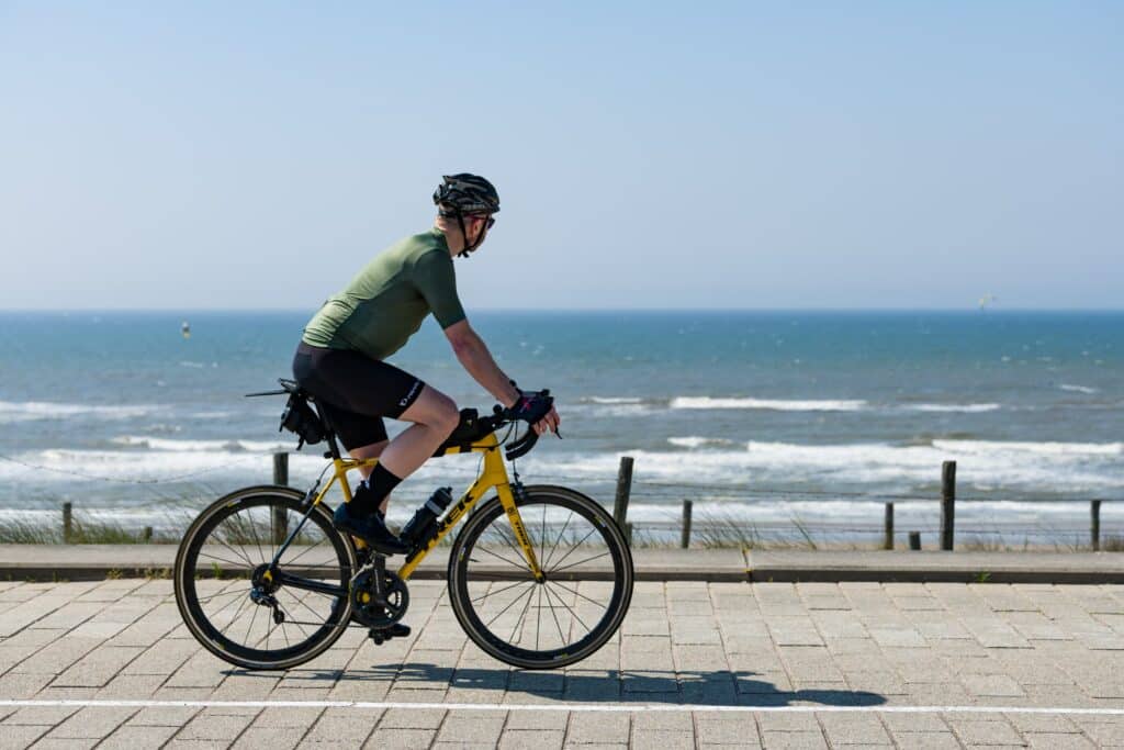 Wielrenner op een gele fiets rijdt over de boulevard langs de zee, met een helm op en kijkt naar de zee.