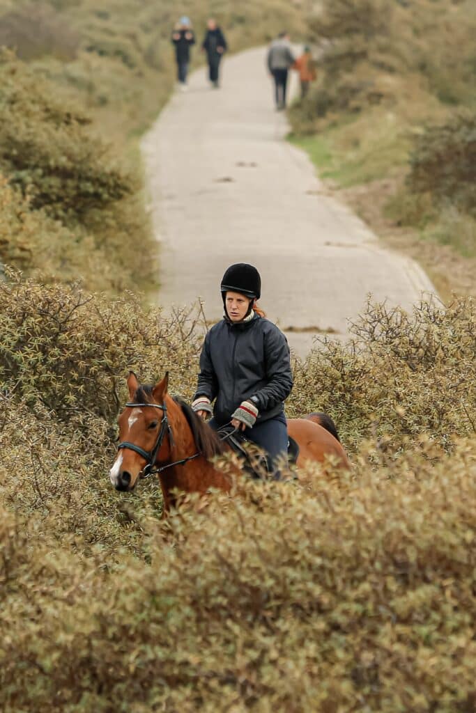 Een vrouw is buiten aan het paardrijden in het Nationaal Park Zuid-Kennemerland, met wandelaars op de achtergrond.