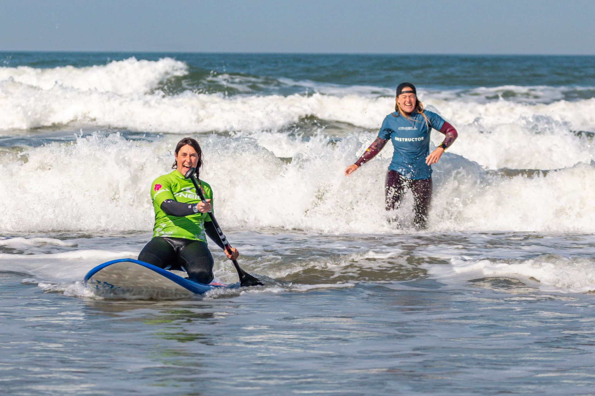 Vrouw die leert suppen zit op haar knieen op een supboard met een peddel in de hand en de instructtrice achter haar staan din de golven