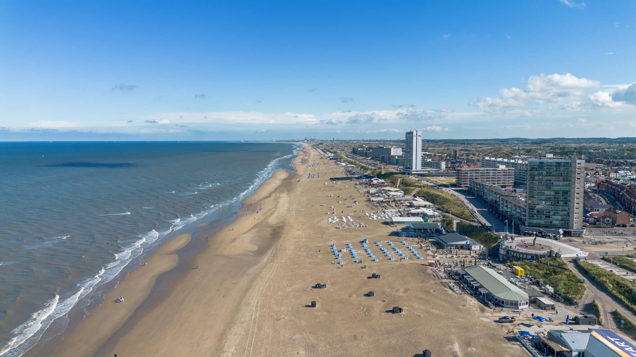Droneoverzicht van het strand, met links de zee, in het midden het strand en rechts het dorp, onder een blauwe lucht op een mooie dag.