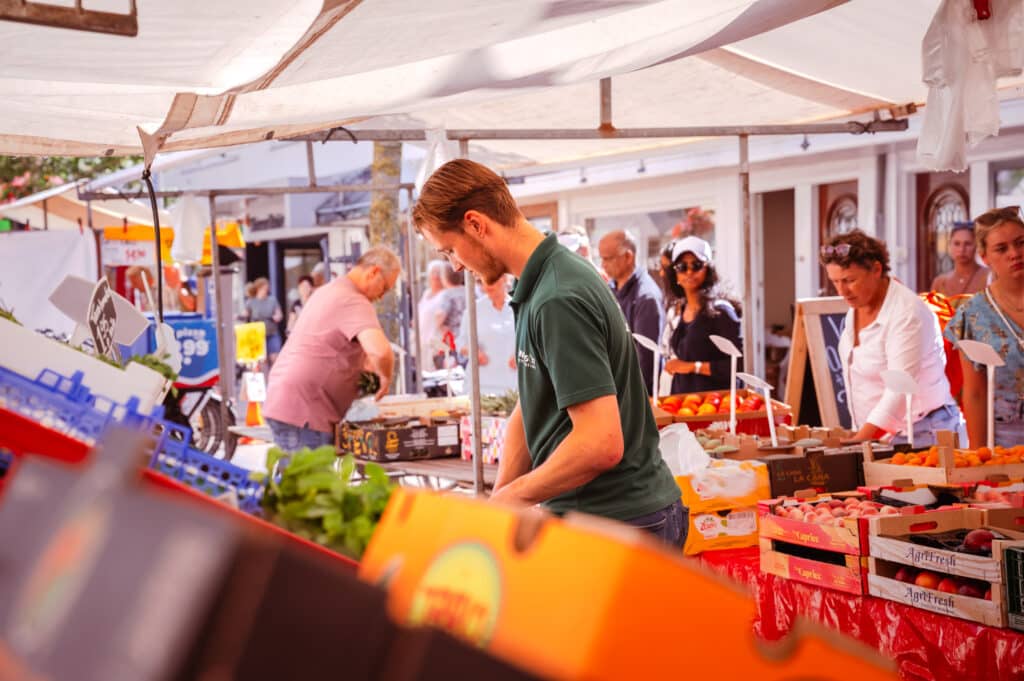De weekmarkt in het centrum van Zandvoort, met een man die werkt bij de groente- en fruitkraam en klanten eromheen.