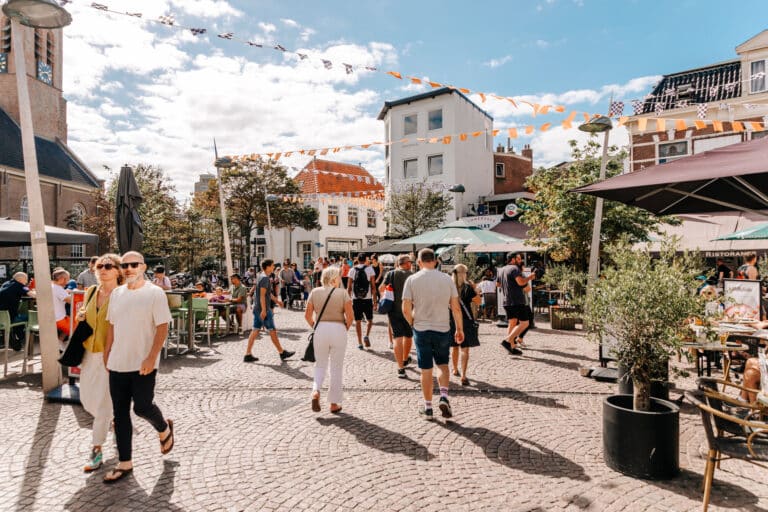 Kerkplein in het dorp Zandvoort, met de kerk op de achtergrond, terrasjes en mensen die op het plein wandelen.