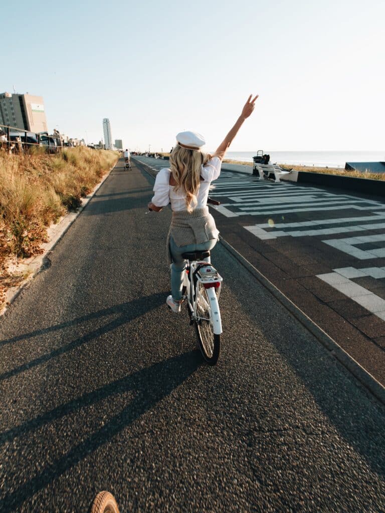 Vrouw fietst langs de boulevard van Zandvoort, met de zee op de achtergrond en haar rechterarm in de lucht.
