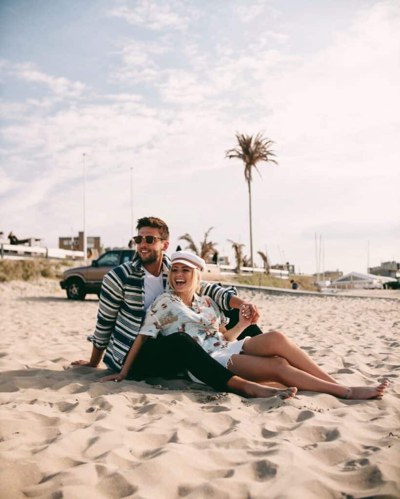 Een lachend stel zit op het strand, terwijl zij tegen hem aanleunt met een palmboom op de achtergrond.