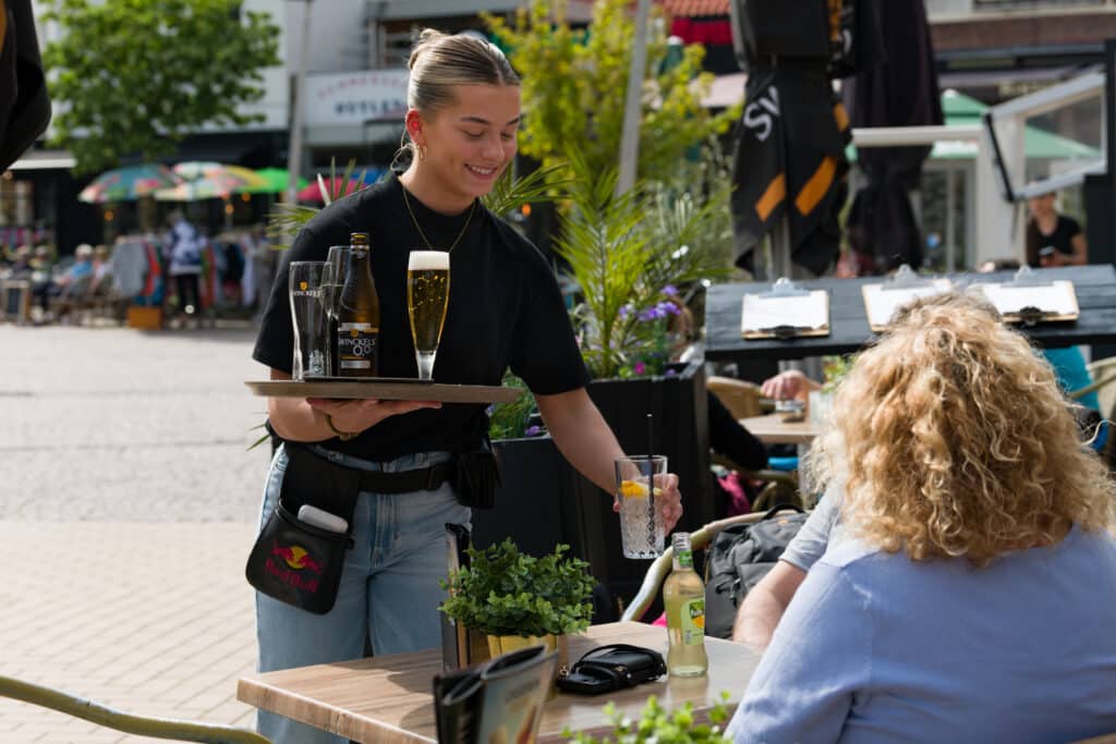 Een vrouw in de bediening serveert drinken aan een stel op het terras op een zonnige dag.