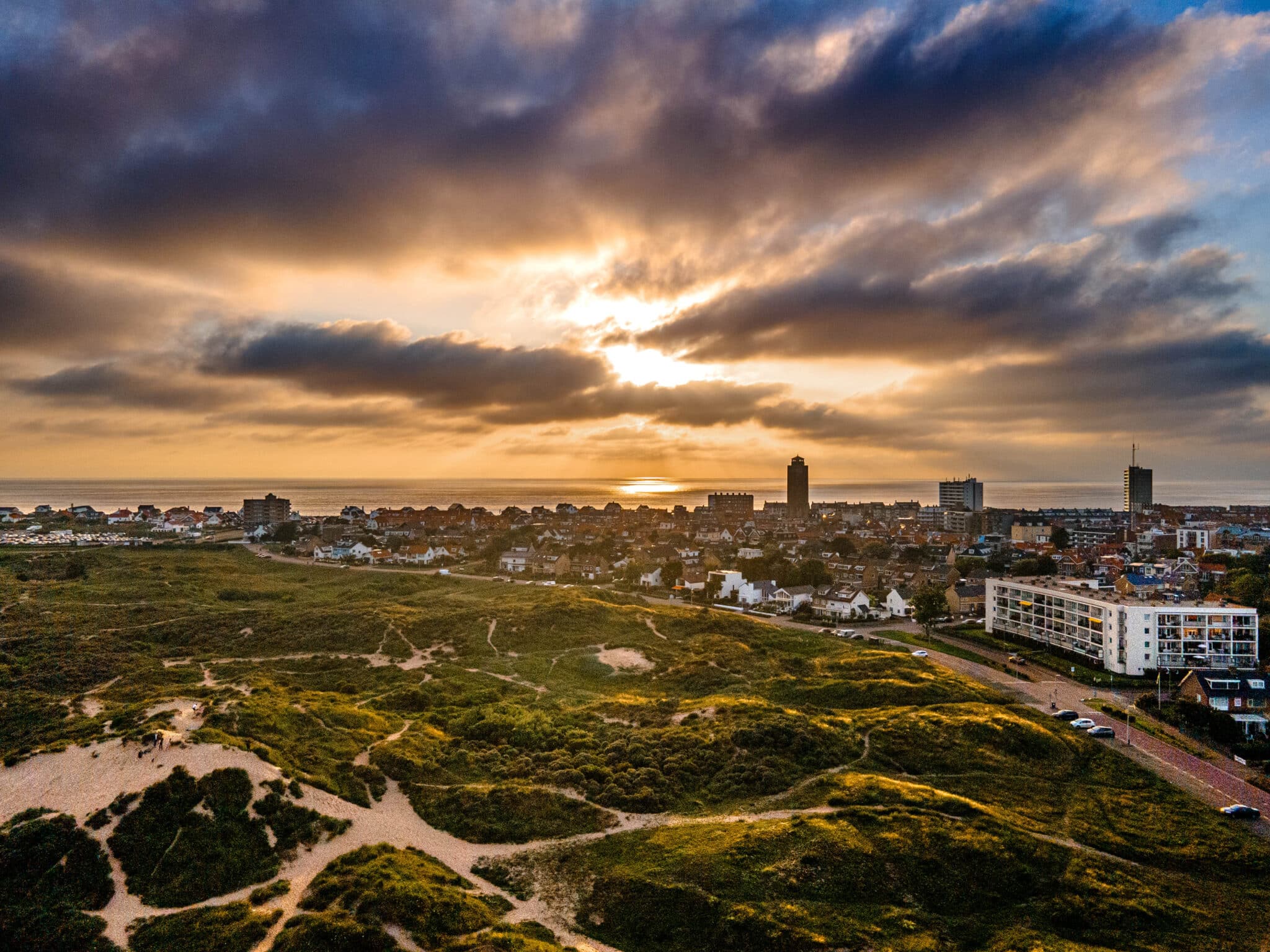 Droneoverzicht van het dorp, met duinen op de voorgrond, huizen en de watertoren op de achtergrond, en de zee in de verte.