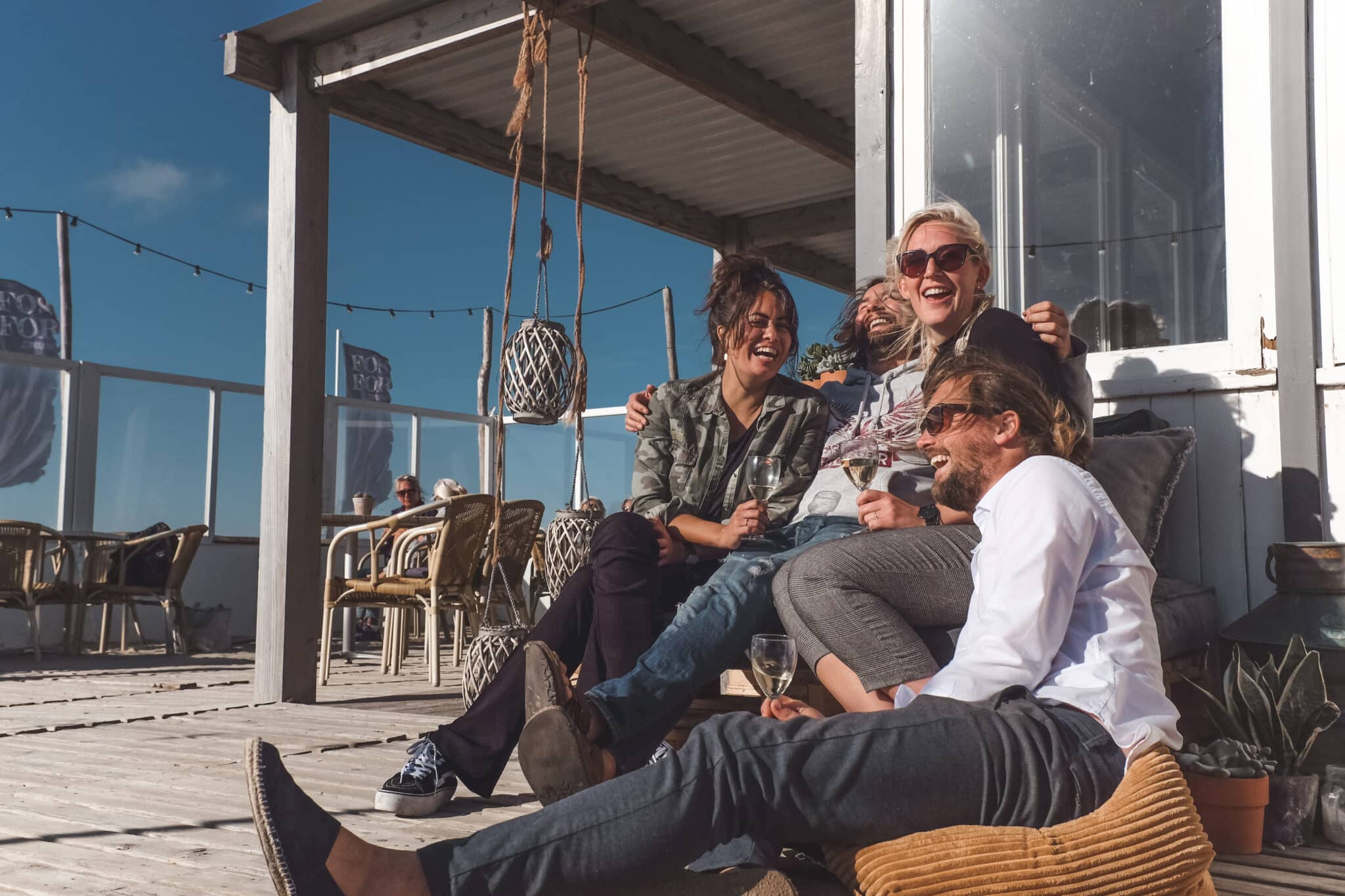 Groep vrienden zit lachend op het terras bij een strandpaviljoens.