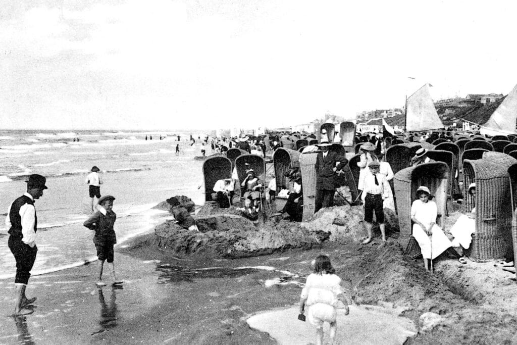 Een historische zwart-witfoto van het strand, met ouderwetse strandstoelen en strandgangers.