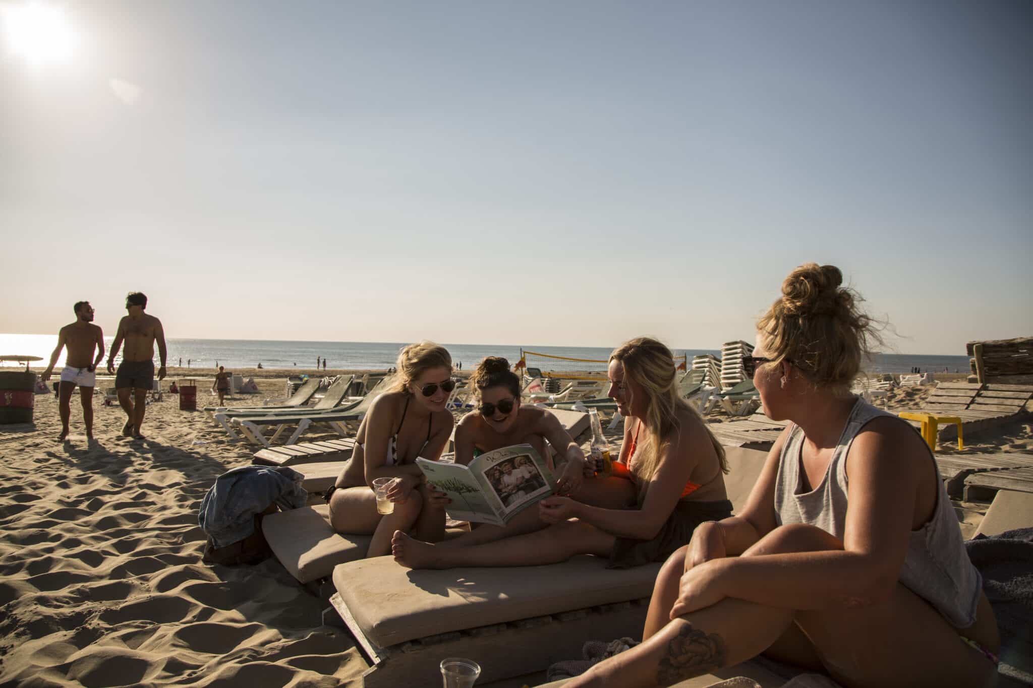 Vier jonge vrouwen op strandbedjes op het strand van Zandvoort bekijken samen een magazine in de ondergaande zon