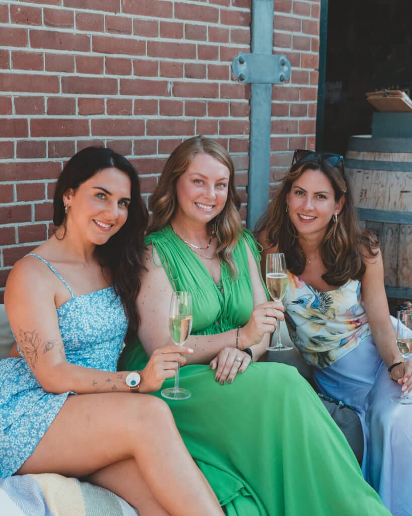 3 vrouwen zitten op een zomerse dag op een bankje voor WIjn aan Zee met een ieder een glas bubbles in hun hand.