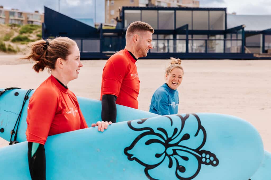 vrouw en man lopen met een surfbord onder de arm samen met de instructrice over het strand