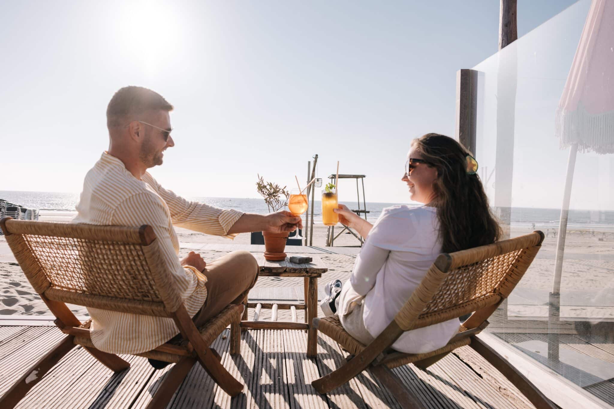 Een koppel op het terras aan het strand op relaxte stoeltjes proostend met cocktails